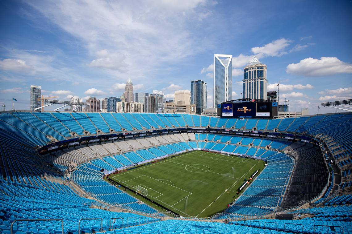 A wide-angle view of an empty football stadium with blue seats and a grass field, set against the backdrop of a city's skyline with modern skyscrapers under a partly cloudy sky.