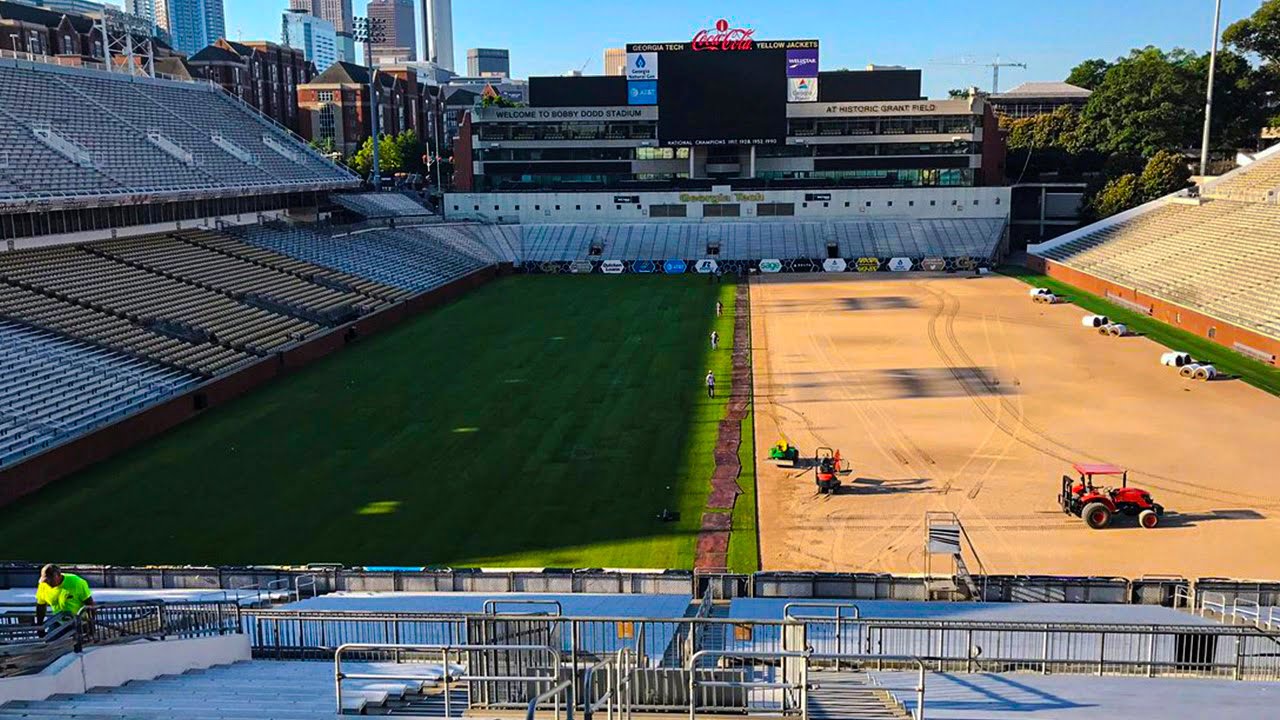 A football stadium under maintenance, with part of the field covered in grass and the other part in dirt. Several workers and tractors are visible. Stands and a scoreboard are seen in the background.