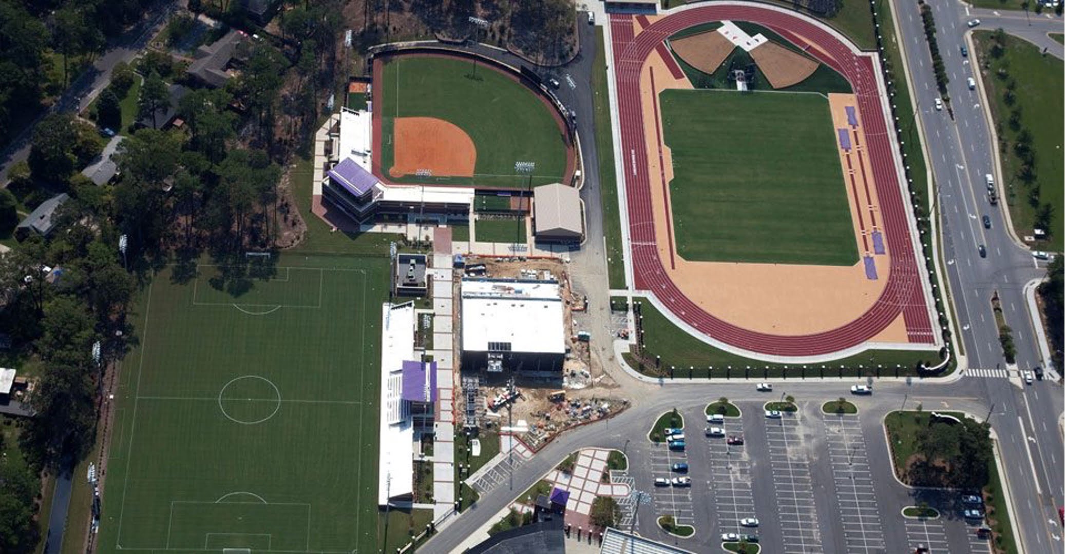 Aerial view of athletic facilities including a soccer field, a baseball field, and a track and field stadium. Indicates ongoing construction near the soccer field. Adjacent roads and parking lots are also visible.