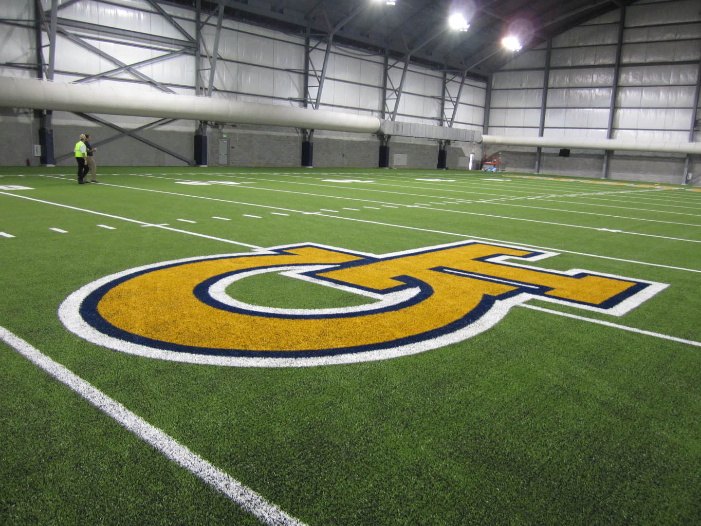 A person stands on an indoor football field with the yellow and blue "GT" logo at the center under a large industrial-style roof with overhead lights.