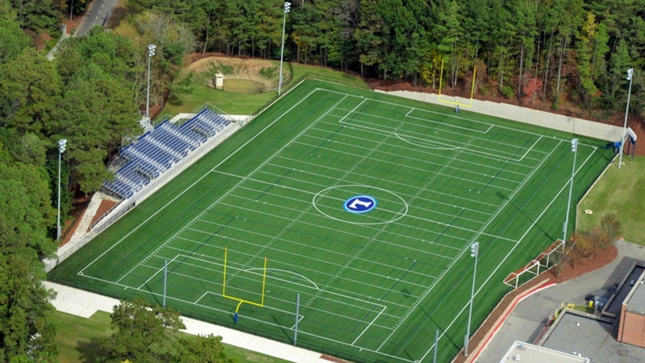 An aerial view of a football field with goalposts, bleachers on one side, surrounded by trees and pathways.