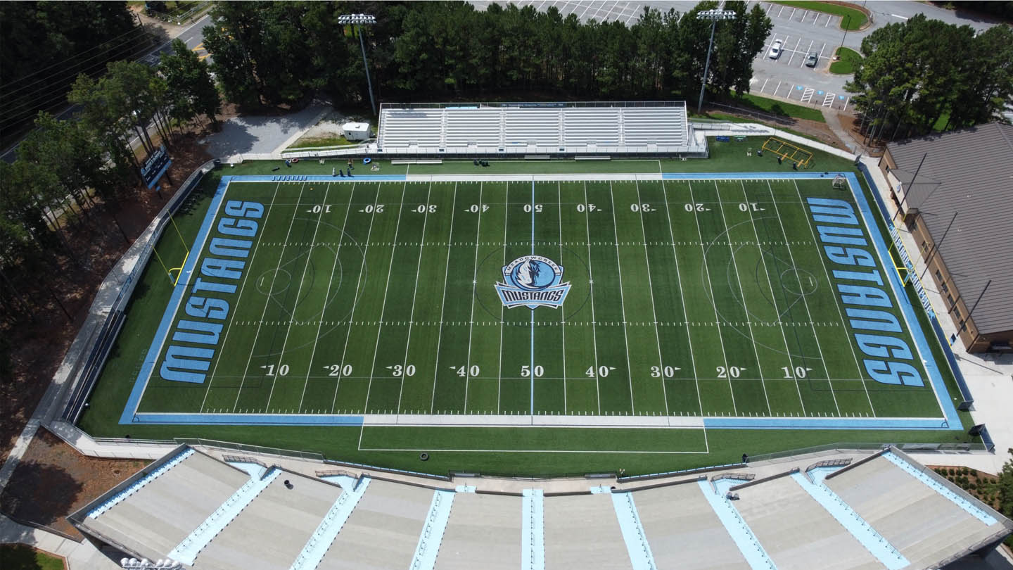 Aerial view of an empty football stadium with "Mustangs" written on both end zones. The field has a central logo, yard lines marked, and surrounding bleachers and lights. Trees and parking lot in the background.