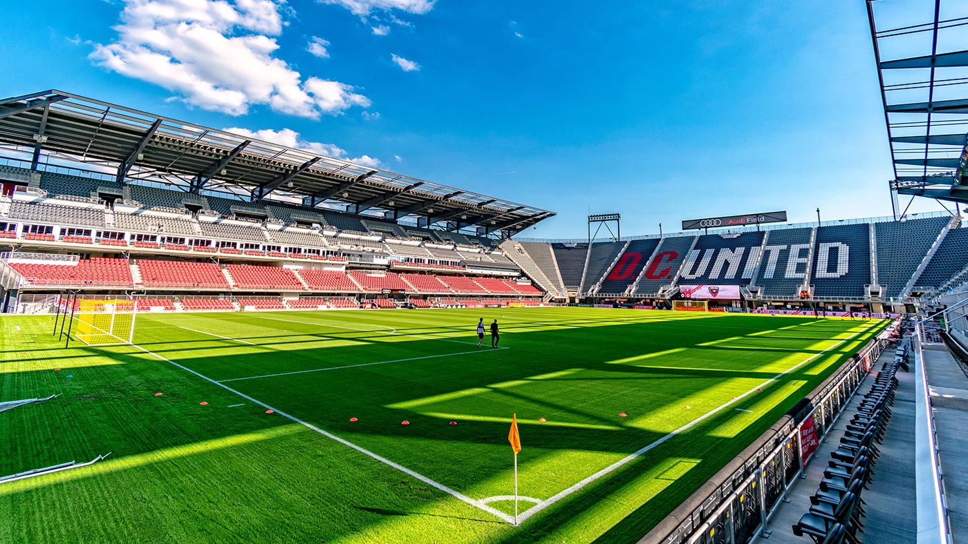 A brightly lit, empty soccer stadium with meticulously maintained sports turf, stands, and a scoreboard displaying "DC United" against a blue sky with scattered clouds.