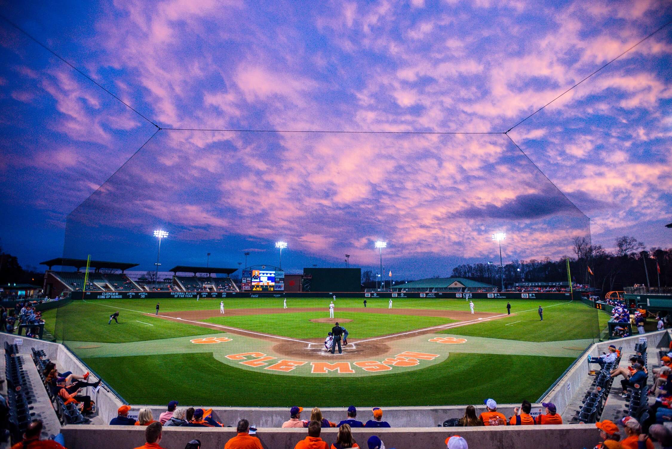 A baseball game in progress at a stadium during sunset, with a vibrant sky of pink and purple clouds. The expertly maintained field, courtesy of a renowned sports turf company, has "Clemson" written behind home plate. Spectators are seated in the stands.