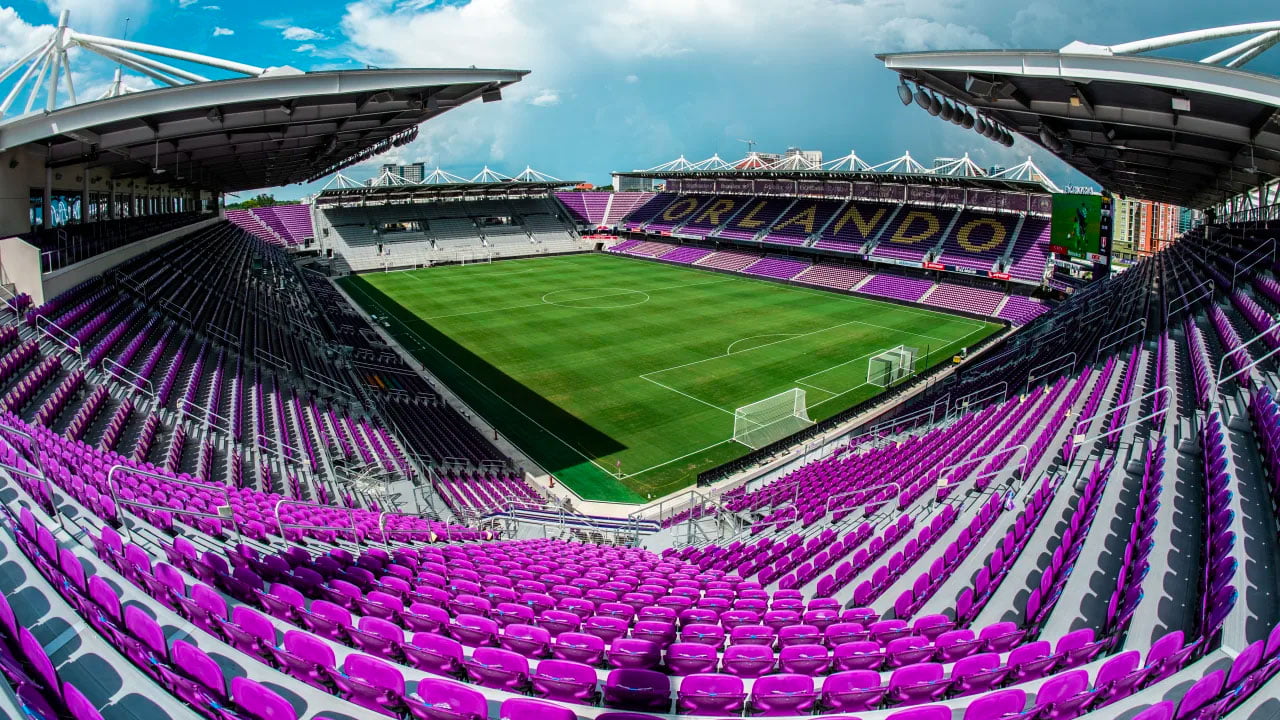 Empty soccer stadium with green field and purple seats. The word "Orlando" is visible on the upper seats. Overhead roof structure covers the seating areas. Bright, partly cloudy sky above.