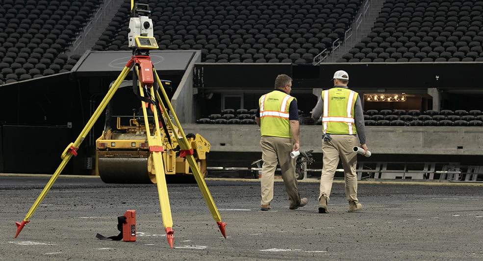 Two construction workers from a sports turf company, wearing safety vests and helmets, walk across a gravel surface in a stadium. Surveying equipment is set up in the foreground.