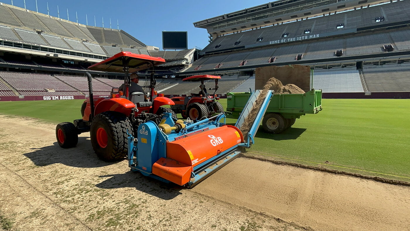 Two tractors are performing precision turf maintenance on a sports stadium field. One is pulling a machine spreading sand from a green trailer, while the other is parked nearby. The stadium is empty.