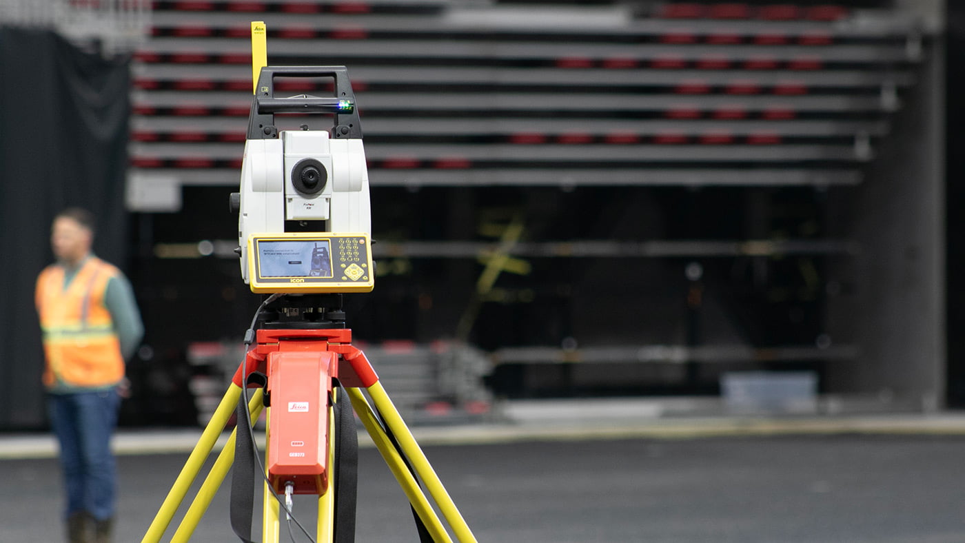 A surveying instrument is set up on a tripod; a man in a high-visibility vest stands in the background, ensuring the precision of their work.