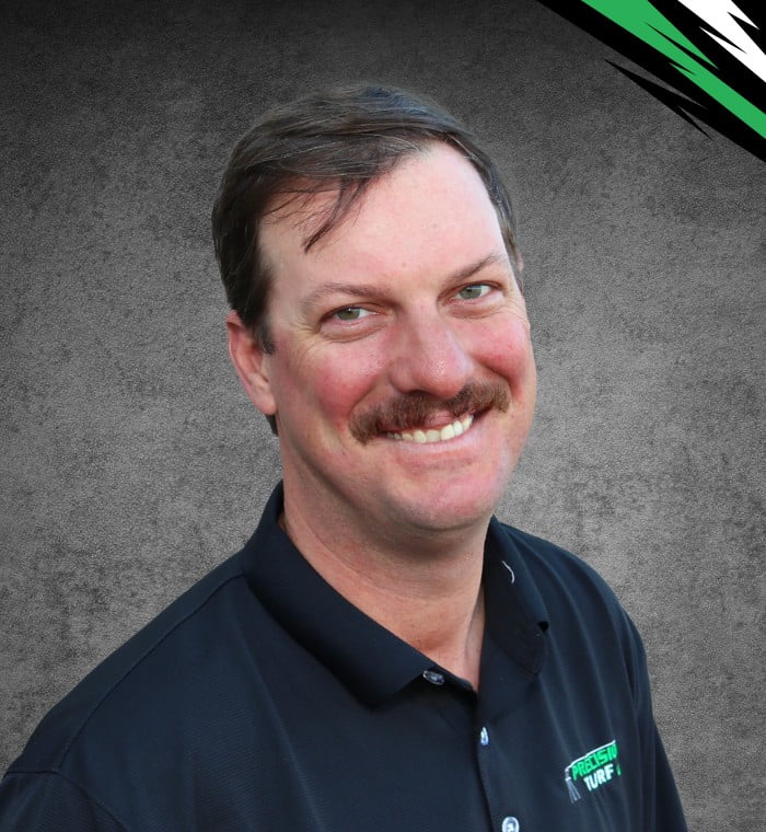 A man with a mustache smiling, wearing a black polo shirt with an embroidered "Turf" logo, stands against a gray background with green, white, and black accents in the corner. This image captures the essence of precision turf management.