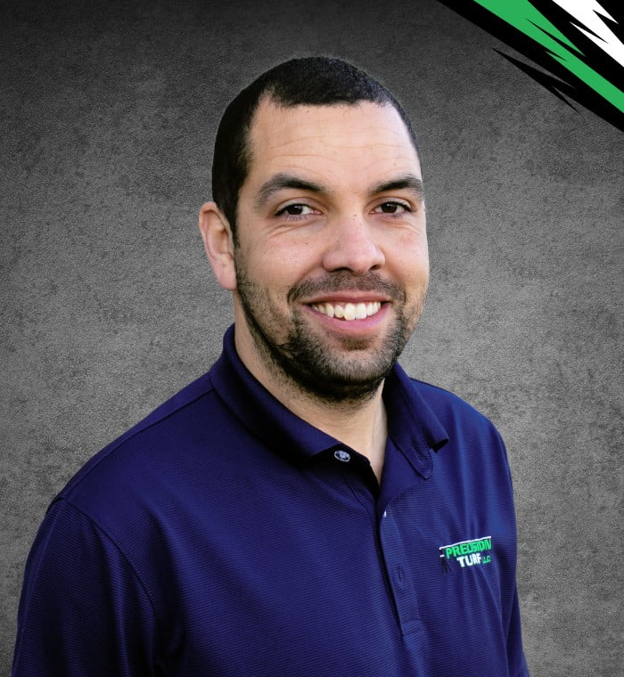 A man with short hair and a beard wearing a navy polo shirt stands against a gray background with a green and black design on the top right corner, looking focused as if he's discussing precision turf.