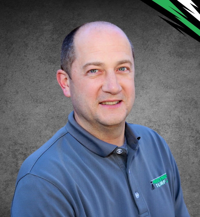 A man wearing a gray collared shirt with a company logo poses in front of a textured gray background, embodying the precision turf expertise he represents.