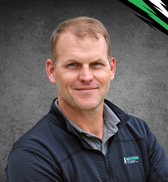 A man with short hair and a slight smile is wearing a gray collared shirt and a black zip-up jacket with a green and white logo that reads "Precision Turf." The background is a gradient of gray.