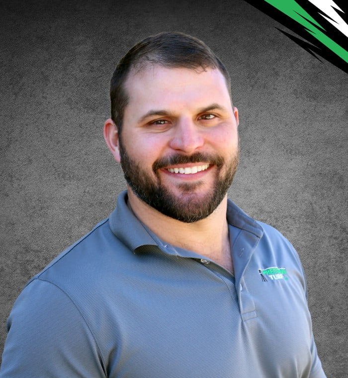 A bearded man with short hair smiles at the camera while wearing a grey, collared shirt. The background features a grey texture and subtly highlights about precision turf with a black and green design in the top right corner.