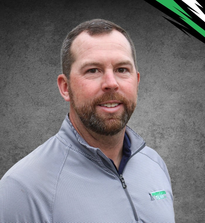 A man with a beard and short hair is wearing a gray zip-up jacket. The background, evoking the precision of shaping turf, has artistic black and green accents in the top right corner.