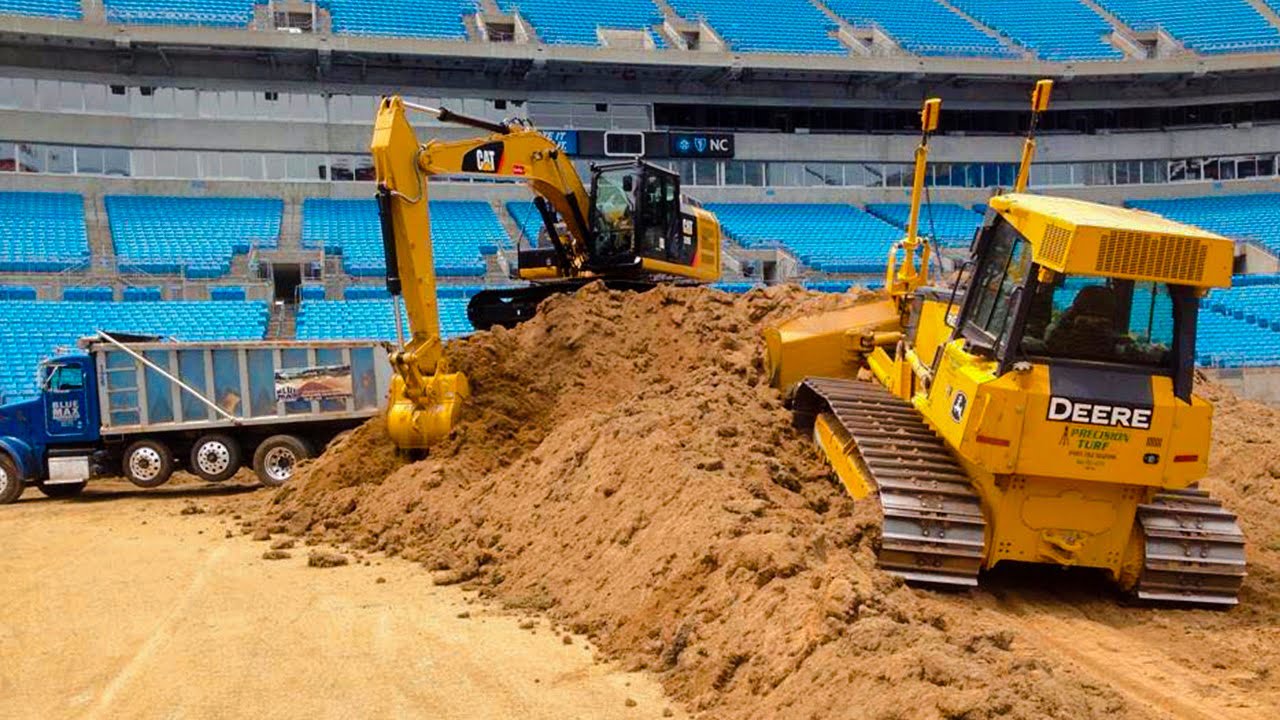 Construction equipment, including a CAT excavator and a Deere bulldozer, expertly moves a large pile of dirt inside an empty stadium with blue seats—preparing the field for precision turf installation. A dump truck is parked nearby.