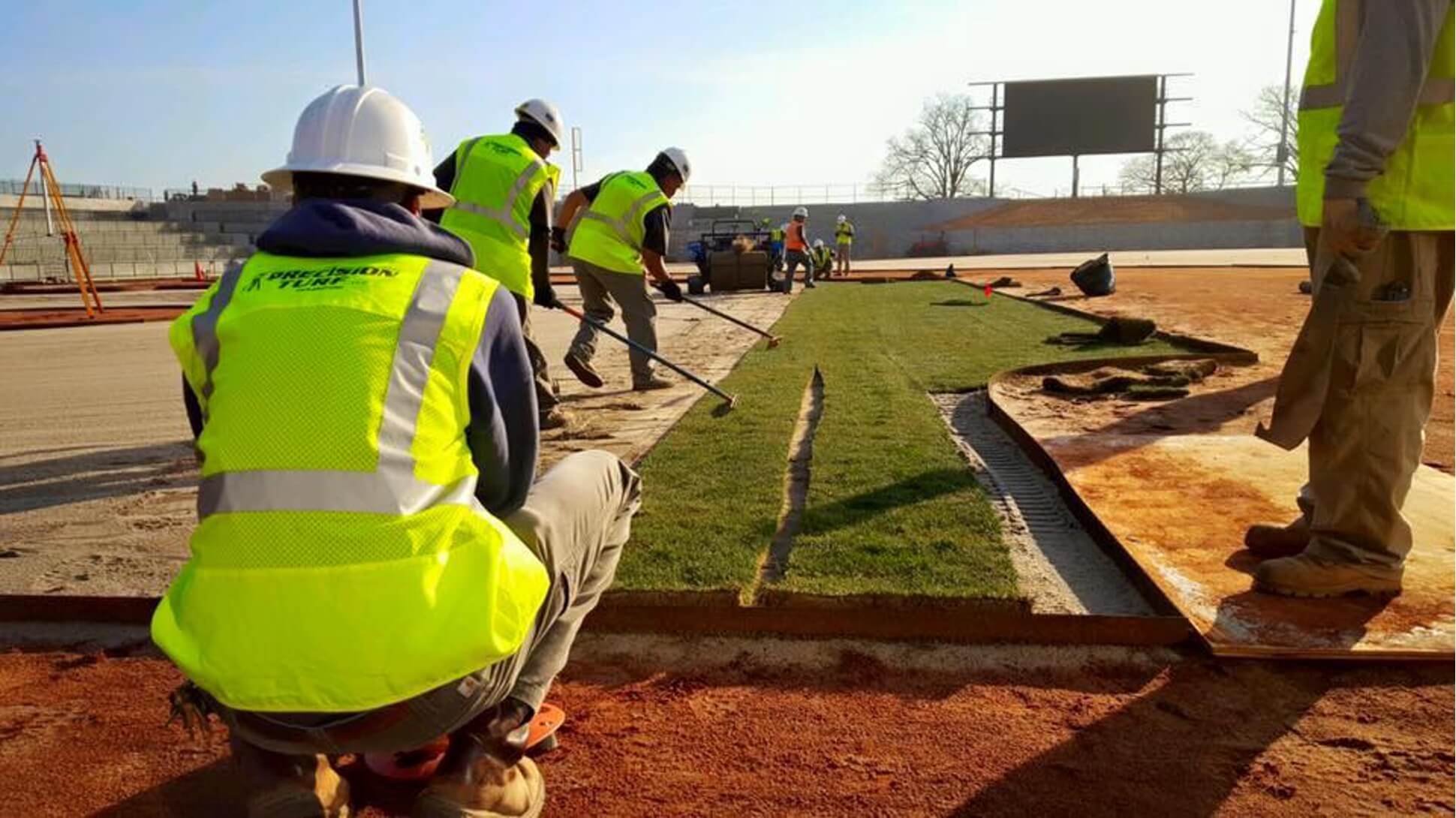 Construction workers in yellow vests and helmets, showcasing their promising careers, install a large piece of turf on a sports field under sunny conditions.