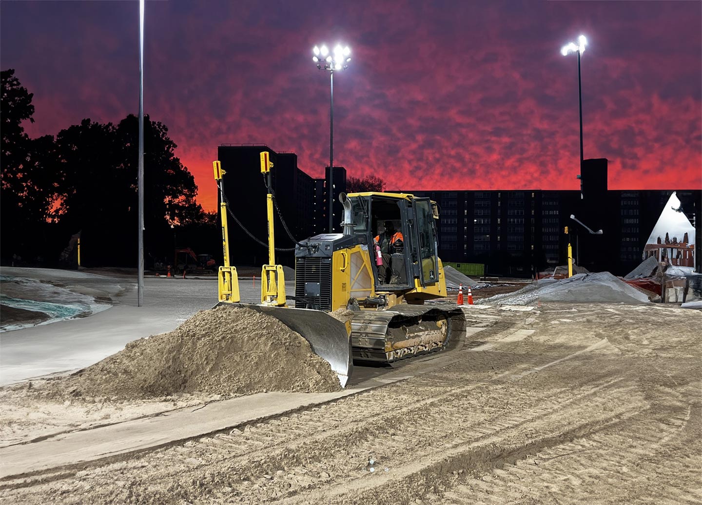 A bulldozer with a worker operates at a construction site during sunset, under the illuminated stadium lights and in front of a darkened building, as part of a project for a renowned sports turf company.