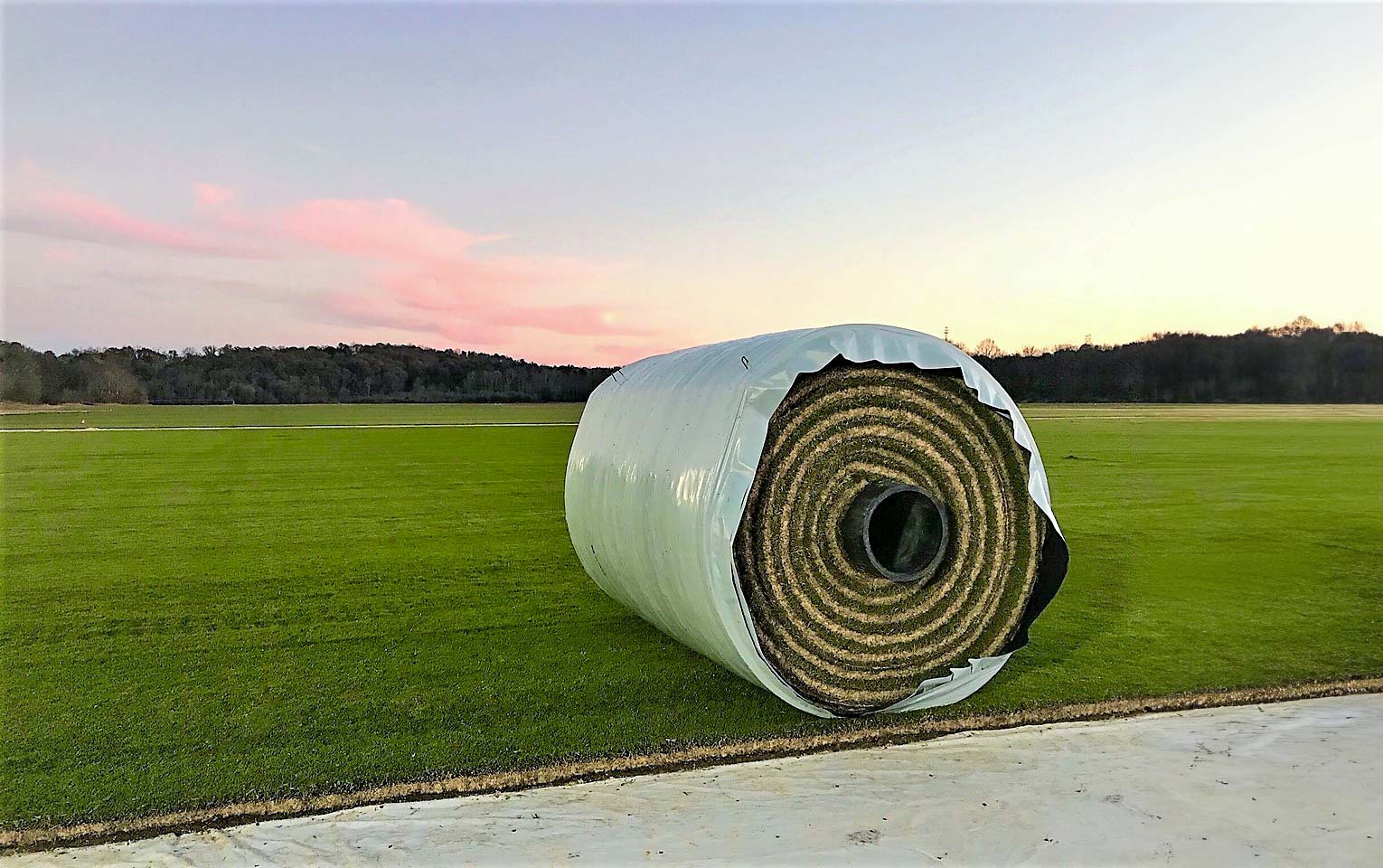 A large roll of instant play turf, wrapped in white plastic, rests on a green field with a tree-lined horizon and a light pink sky.