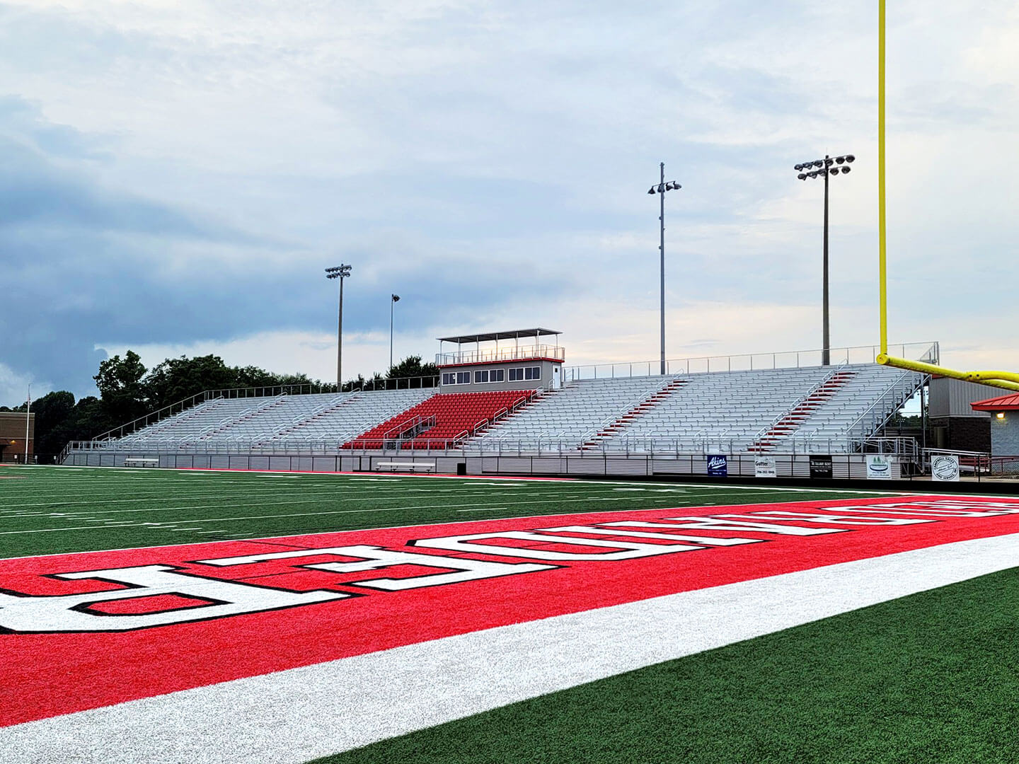 An expertly planned sports field design showcases an empty football field with a red end zone and a vast, unoccupied seating area, all set beneath a cloudy sky.