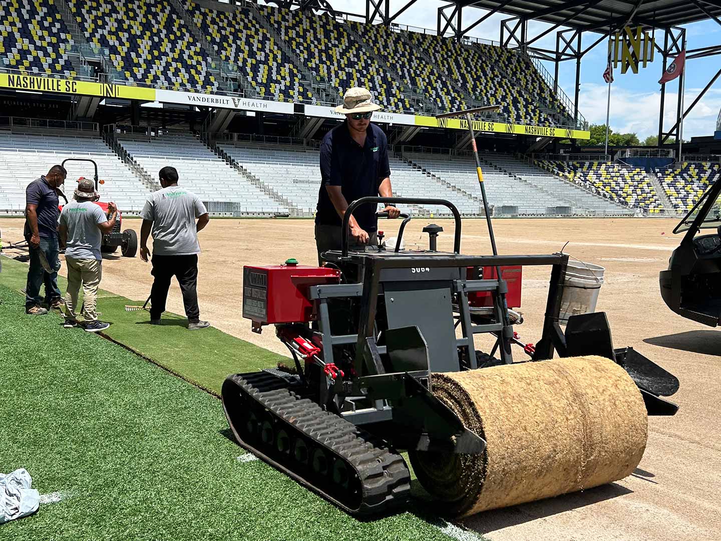 A worker operates a machine to install instant play turf on a sports field, while other workers assist in the background. The stadium seats are visible, with some covered in yellow and black designs.