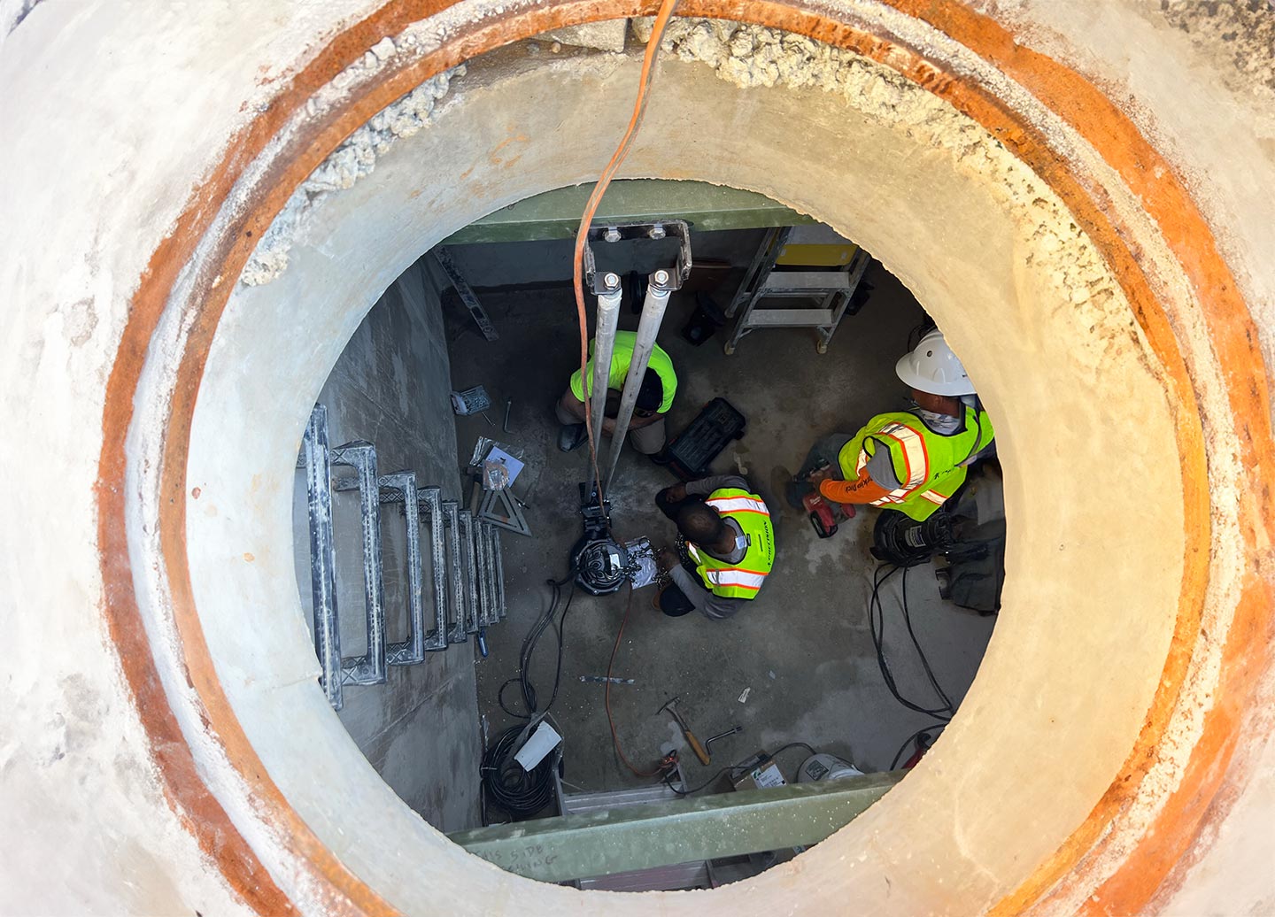 View from above shows three workers in safety gear maintaining a large, circular concrete structure with tools and equipment, surrounded by natural turf.