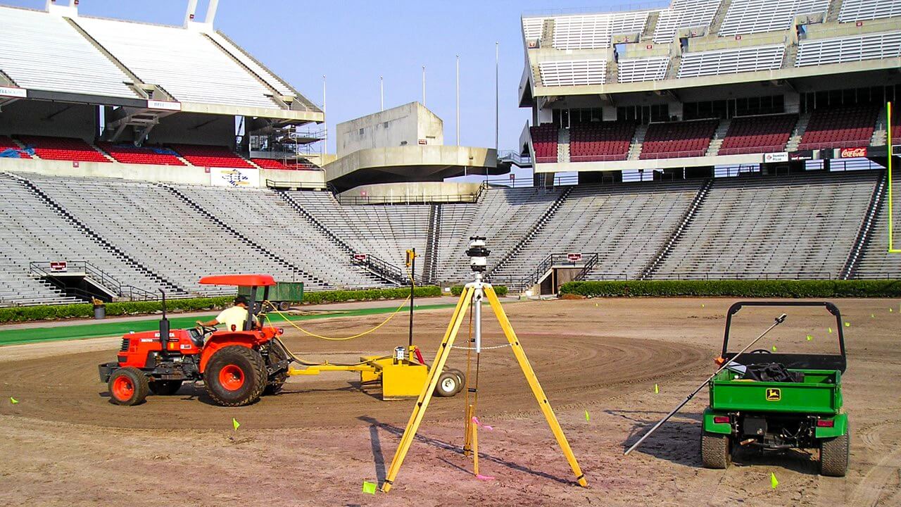 Workers on tractors and utility vehicles prepare the ground at a large stadium, with seating visible in the background and surveying equipment set up at the center, showcasing meticulous sports field design.