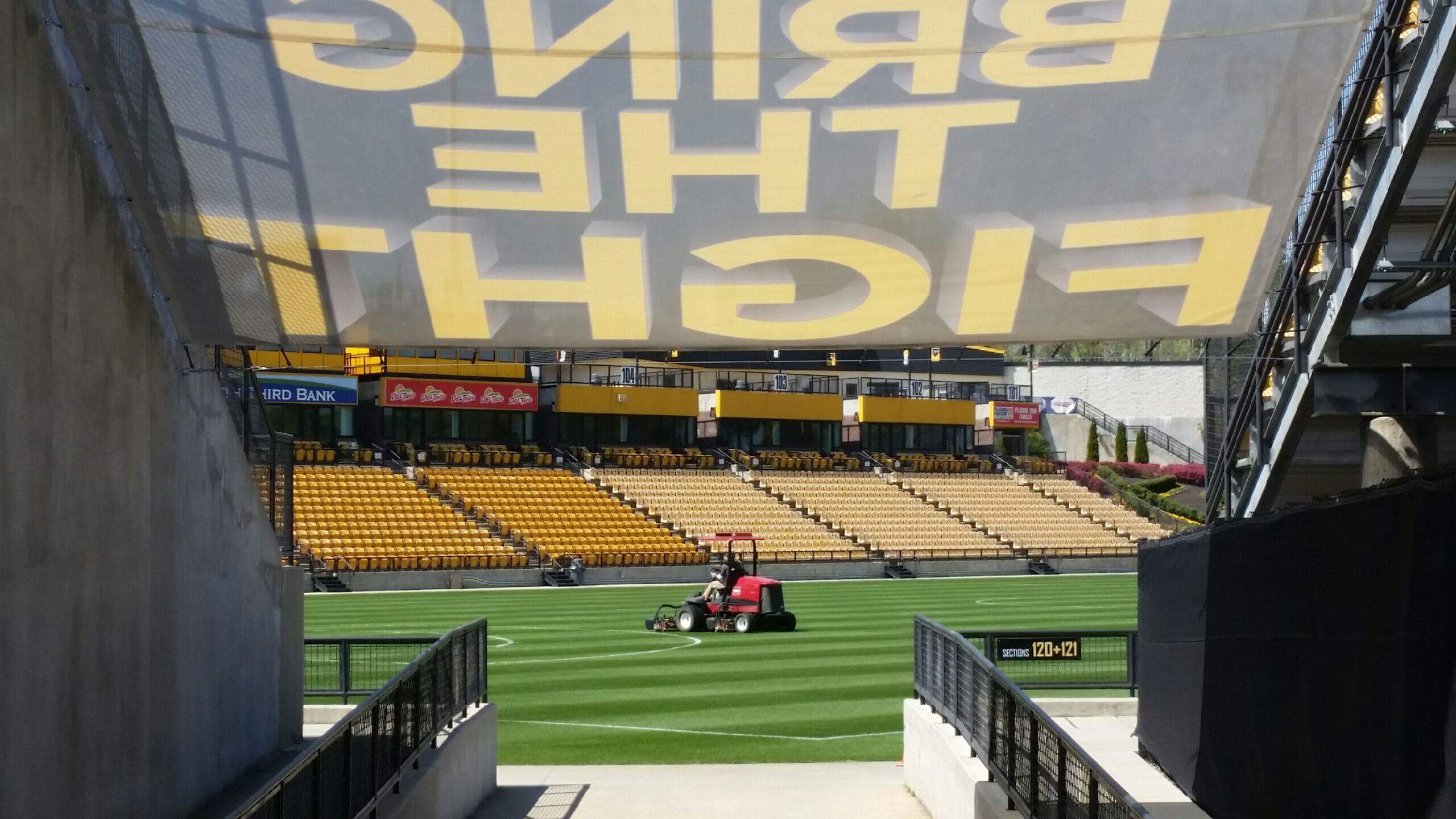 View from a concourse tunnel leading to the seating area in a stadium, showing a groundskeeper mowing the natural turf, with an overhead banner displaying "BRING THE FIGHT" in large yellow letters.