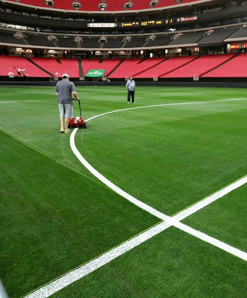 Two workers are painting white lines on the natural turf of a football field inside a large, empty stadium.