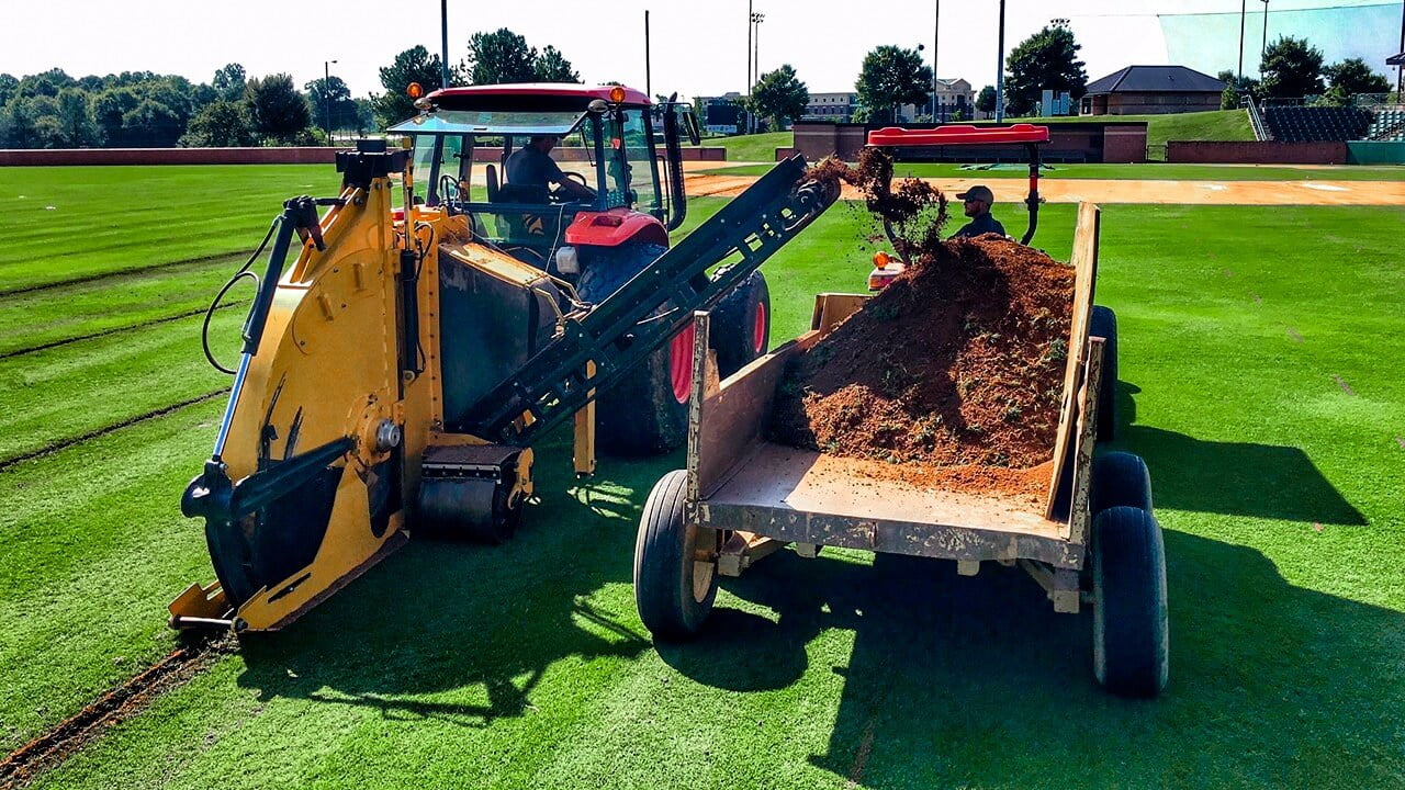 Two tractors are working on a green field; one is digging a trench while the other is collecting the dug-up soil, precisely shaping the land for an upcoming sports field design.