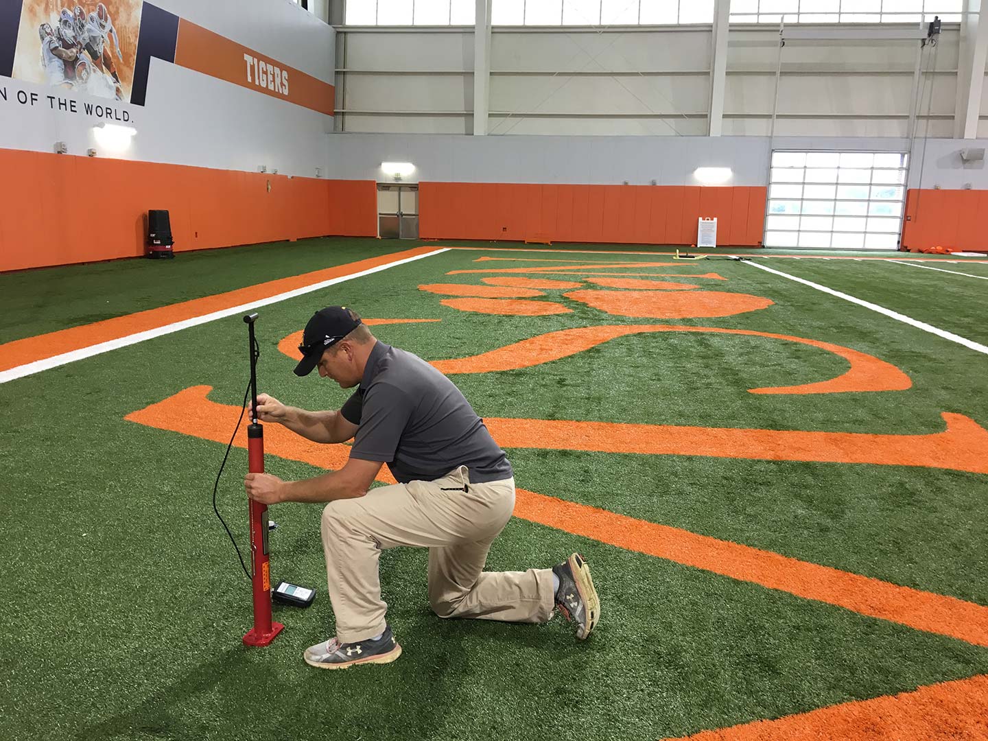 Man kneels on synthetic turf football field, using equipment placed into the ground, inside an indoor sports facility with orange and white colors.